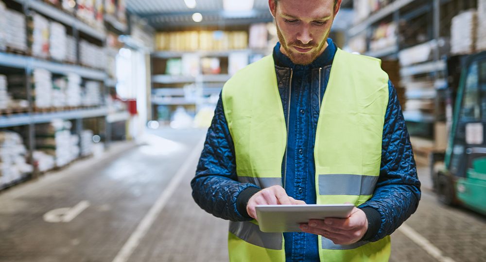 A bearded man wearing a high-vis jacket using a tablet in a warehouse