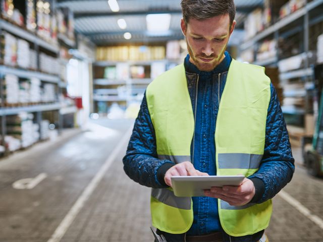 A bearded man wearing a high-vis jacket using a tablet in a warehouse